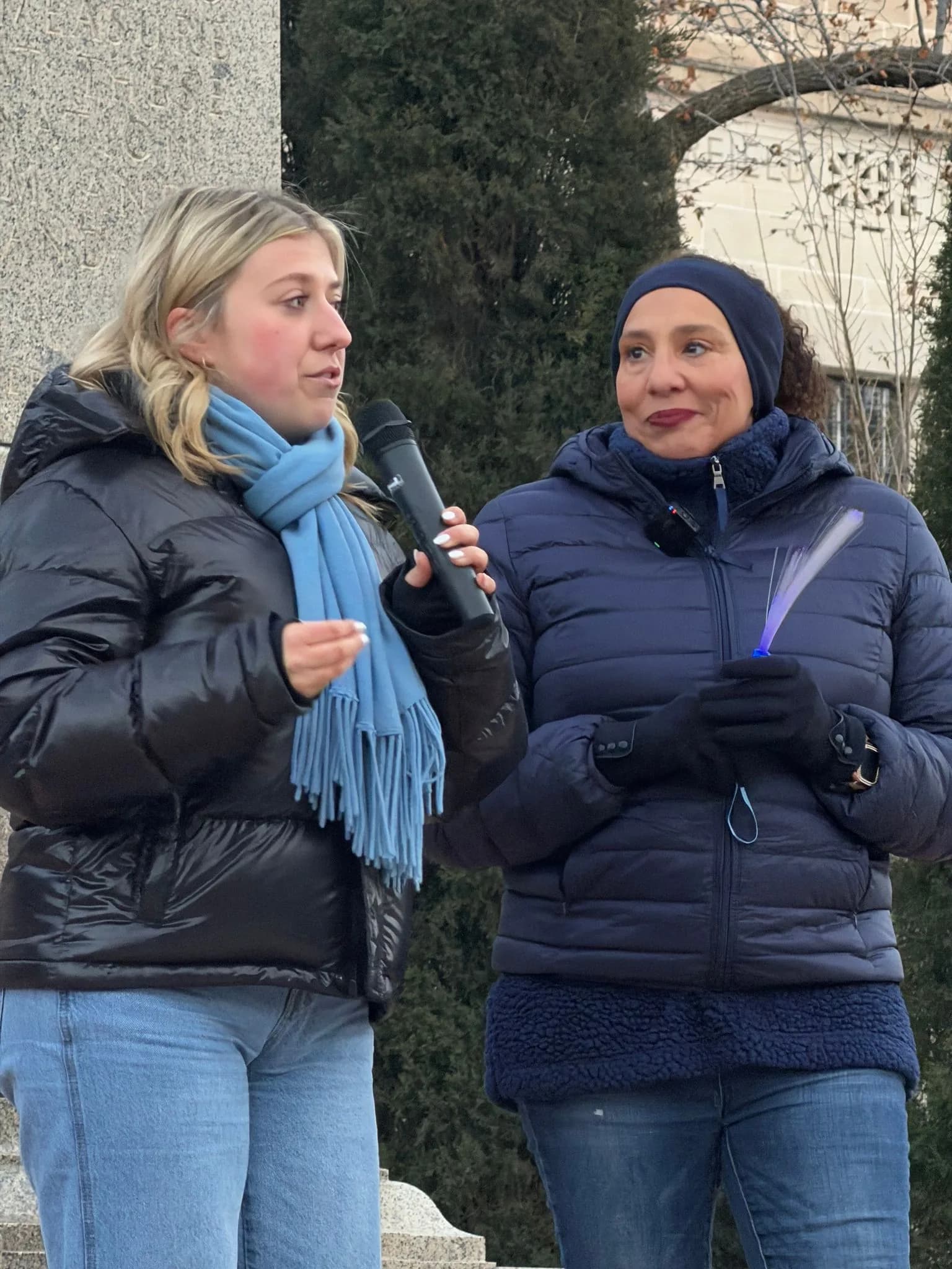 Two women in winter coats, one speaking into a microphone and one holding a glow stick