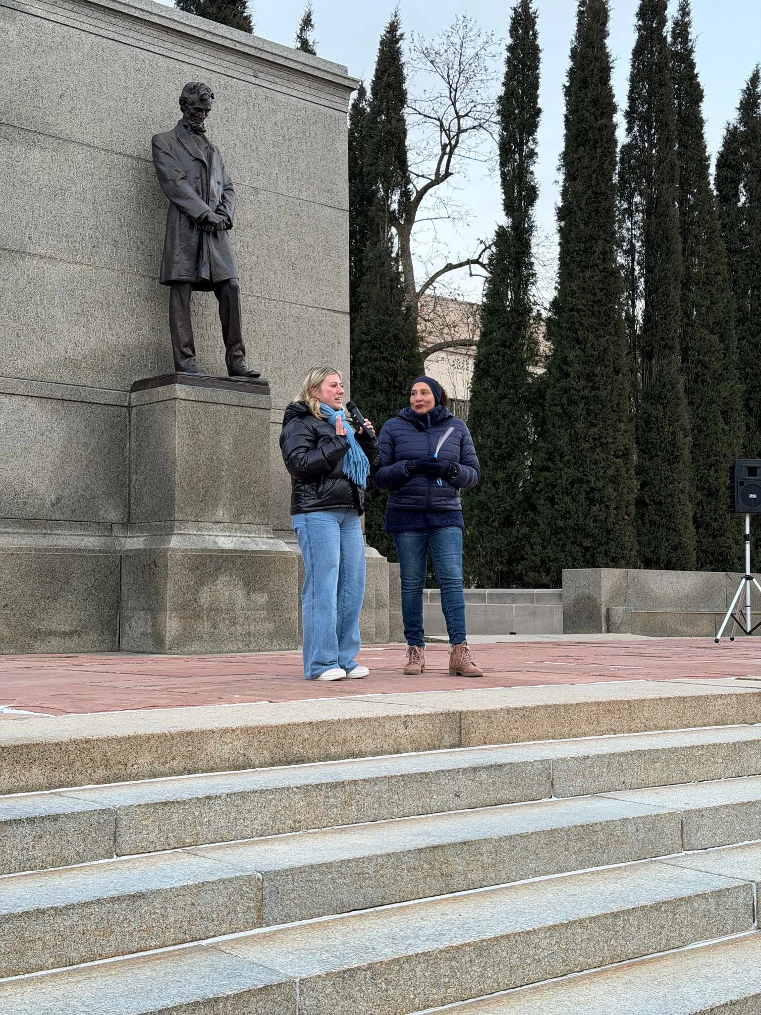Two speakers on a stone plaza below a statue on a tall plinth
