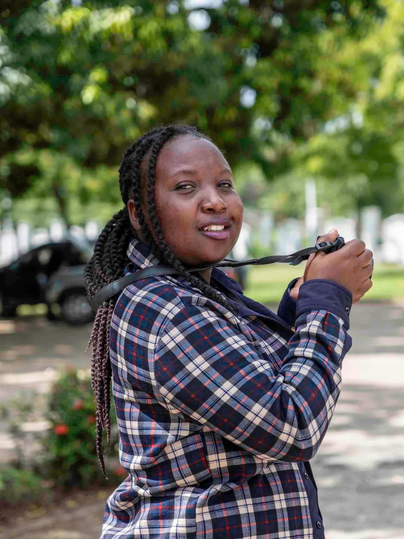 A smiling woman in a plaid shirt holding a camera by its strap outdoors
