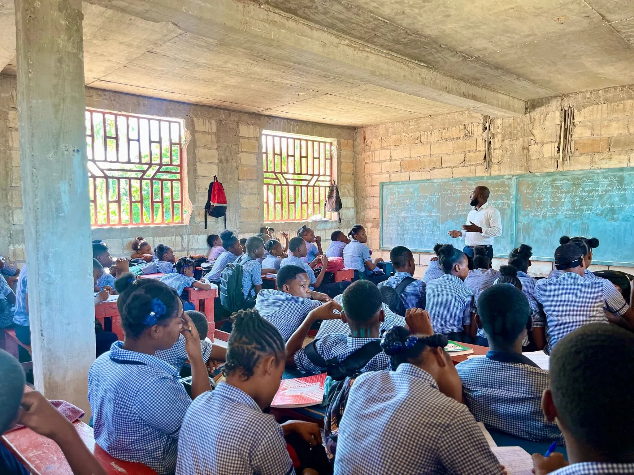 A full high school classroom seen from the back, students in blue uniforms facing a teacher