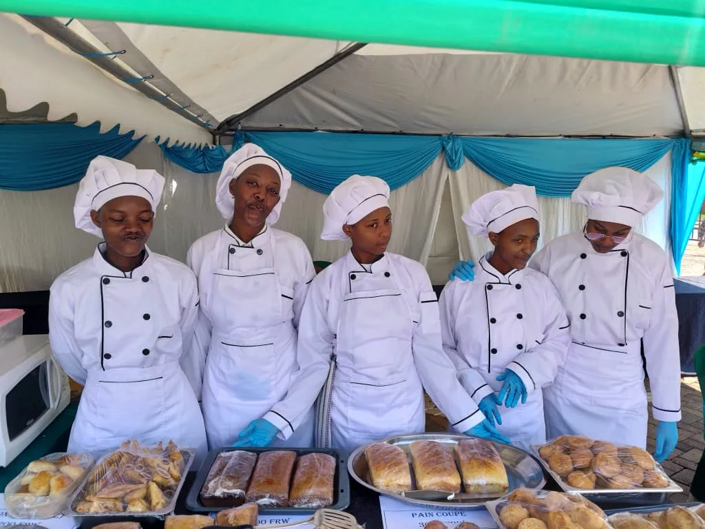 Students in chef whites and hats behind a table of bread and pastries under a tent